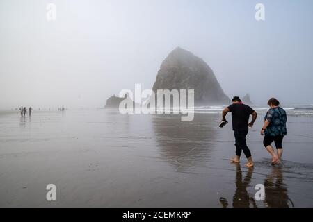 Cannon Beach, Oregon - 1er août 2020 : couple touristique marche pieds nus dans l'océan près de Haystack Rock Banque D'Images
