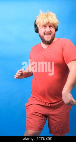 Drôle de chubby freak homme dans le casque et une perruque dans un t-shirt rose danse et rires sur un fond bleu, vertical Banque D'Images