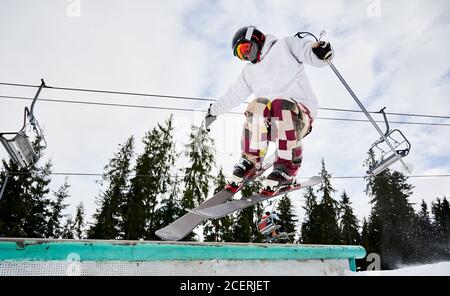 Skieur mâle dans une veste de ski et un casque faisant sauter le jour d'hiver. Homme freerider saut dans les airs à la station de ski avec des pins et des remontées mécaniques en arrière-plan. Concept de sports d'hiver et de loisirs actifs. Banque D'Images