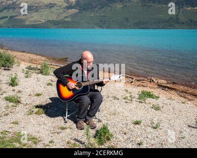 Un bald blanc d'âge moyen jouant une guitare acoustique À l'extérieur, sur la rive du lac Wakatipu, en Nouvelle-Zélande Banque D'Images
