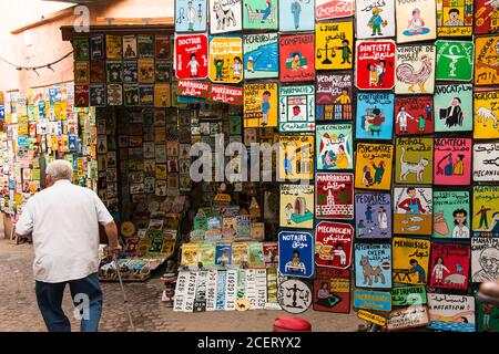 Un homme avec un bâton de marche passe devant un stand vendant des panneaux peints à la main dans le souk dans la médina, Marrakech Banque D'Images