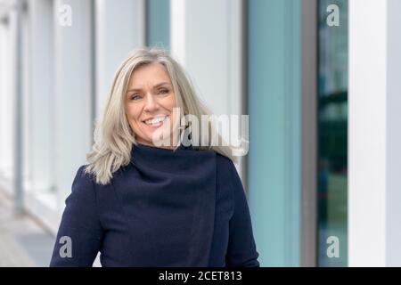 Femme blonde d'âge moyen, confiante et élégante, qui se promette dans une ville rue avec la brise soufflant ses cheveux longs comme elle sourit à l'appareil photo Banque D'Images