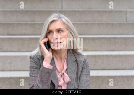 Belle femme d'âge moyen de l'écoute d'un appel sur un mobile tout en étant assis sur des mesures concrètes à l'extérieur de la ville Banque D'Images