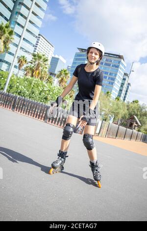 Jeune patineuse à roulettes heureuse Femme caucasienne dans le casque blanc et noir vêtements sportifs, jour ensoleillé, parc à roulettes, environnement urbain. Composition diagonale. Banque D'Images