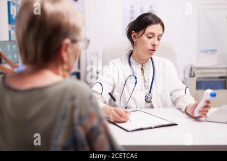 Medic écrit un diagnostic de patient senior après avoir mesuré la température corporelle avec un thermomètre dans le bureau de l'hôpital. Médecin avec stéthoscope portant une couche blanche. Banque D'Images