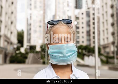 Mignon adorable petite caucasienne bonne blonde fille fermé les yeux dedans lunettes de soleil portant un masque facial de protection en raison de la quarantaine et de la marche de covid-19 en ville Banque D'Images