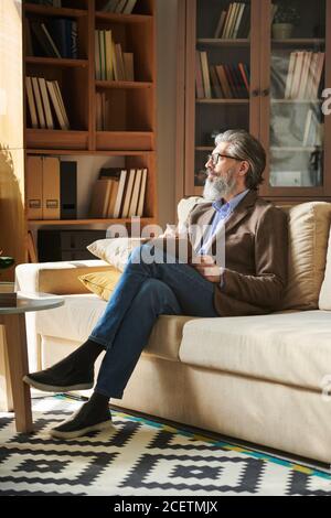Photo verticale d'un bel homme barbu avec une tenue élégante lunettes assises sur un canapé dans une salle de bureau moderne tenant une planchette à pince vue sur l'extérieur Banque D'Images