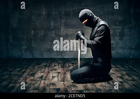 Homme guerrier assis sur le sol posant avec une épée dessus arrière-plan sombre Banque D'Images