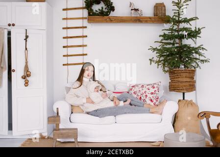 Une jeune mère avec un enfant d'un an dans ses bras est assise sur un canapé blanc dans le salon, décoré pour Noël et le nouvel an Banque D'Images