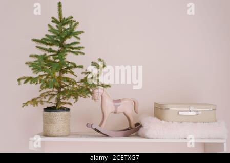 Un sapin de Noël dans un pot en osier se dresse sur une étagère, à côté d'un cheval à bascule en bois, d'une petite valise et d'une couverture. Décor pour Noël et Banque D'Images