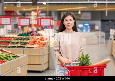 Belle jeune femme adulte portant des vêtements décontractés debout avec des achats chariot dans l'allée du magasin Banque D'Images