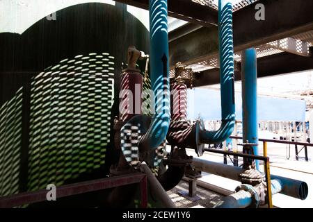 Réservoir en métal, tuyaux et raccords sur l'ombre du sol en maille. Vue rapprochée abstraite de l'usine chimique. Banque D'Images