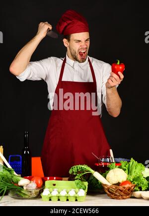 L'homme en uniforme apporte le couteau au poivre. Chef au visage furieux tient le couteau et le poivron rouge sur fond noir. Cuisiner travaille dans la cuisine près des légumes et des outils. Ustensiles de cuisine et concept de cuisine. Banque D'Images