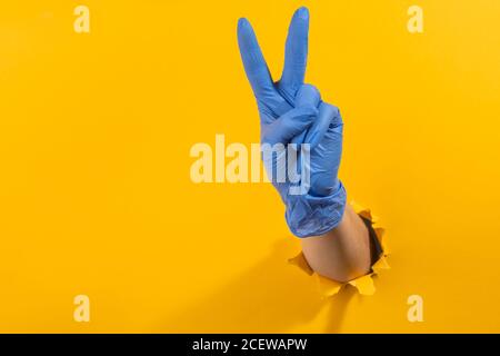 Main dans un gant en latex montrant un signe de victoire mur jaune déchiré Banque D'Images