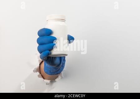 Main dans un gant en latex tenant une bouteille en plastique de pilules à travers le mur blanc déchiré Banque D'Images