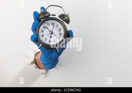 Main dans un gant en latex tenant un réveil vintage à travers un mur blanc déchiré Banque D'Images