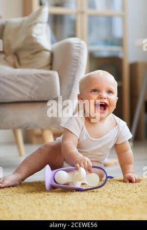 Photo verticale d'un bébé joyeux et amusant assis sur le sol dans le salon, jouer avec le jouet et rire Banque D'Images