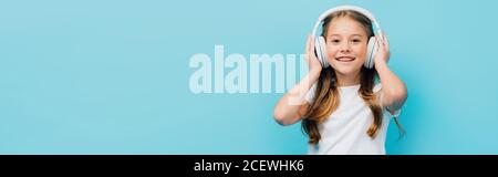 image horizontale de l'enfant dans un t-shirt blanc touchant un casque sans fil isolé sur bleu Banque D'Images