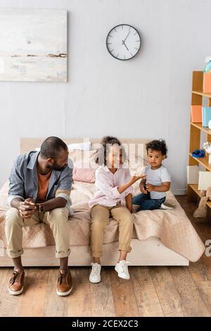 jeunes afro-américains père et enfants dans des vêtements décontractés assis sur le lit à la maison