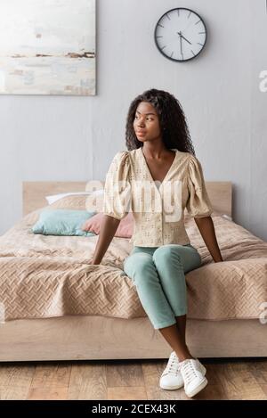 femme afro-américaine pensive en chemisier et pantalon assis sur lit et vue Banque D'Images