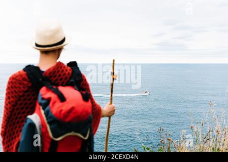 Vue arrière d'une randonneur anonyme avec sac à dos et bâton debout sur une colline et en admirant la mer ondulée avec un bateau flottant Tout en se reposant pendant la route de Camino de Santiago en Espagne Banque D'Images