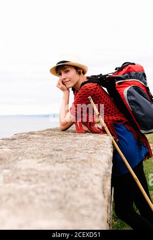 Vue latérale de jeunes femmes pèlerin avec sac à dos et bâton de trekking debout sur la côte de mer regardant la caméra pendant Voyage le long de Camino de Santiago en Espagne Banque D'Images