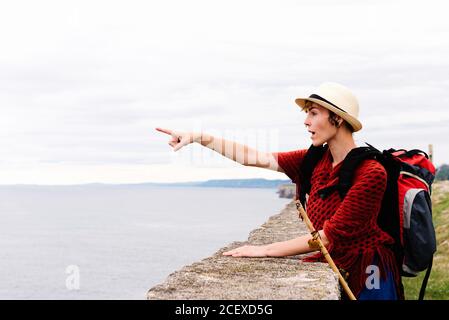 Vue latérale de jeunes femmes pèlerin avec sac à dos et bâton de trekking debout sur la côte de mer et pointant vers l'extérieur pendant Voyage le long de Camino de Santiago en Espagne Banque D'Images