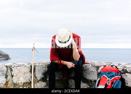 Une femme perdue triste et méconnaissable avec un bâton de randonnée la frontière en pierre près de la mer et la navigation de smartphone tout en se reposant et Vérification de l'itinéraire lors du pèlerinage à travers l'Espagne Banque D'Images