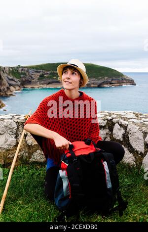 Corps complet de routard jeune femme heureux dans des vêtements élégants assis près de la frontière de pierre contre la baie de mer et regardant loin Tout en se reposant pendant la route de Camino de Santiago en Espagne Banque D'Images