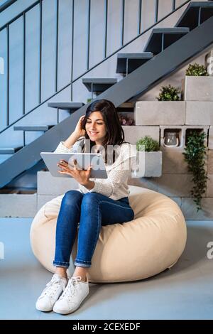 Pleine longueur de jeune femme designer ciblée dans des vêtements décontractés assis sur un repose-pied confortable et utilisant une tablette graphique pendant le travail dans un espace de travail créatif contemporain Banque D'Images