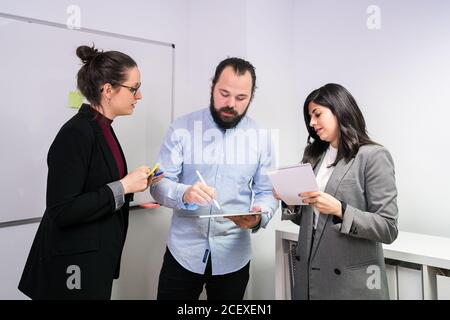 Groupe de jeunes collègues positifs dans les vêtements formels de rassemblement dans bureau de travail de collègue contemporain avec tablette en pause pendant le travail Banque D'Images