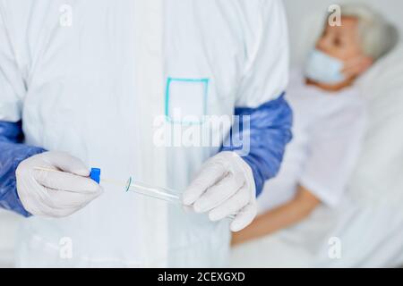 Médecin méconnaissable en uniforme mettant le bâtonnet d'échantillon d'écouvillon dans le test tube après l'avoir pris d'une femme sénichée sur le lit derrière lui Banque D'Images