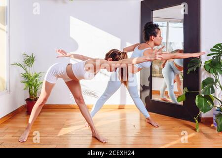 Vue latérale des femelles flexibles en forme d'activewear debout à Trikonasana avec bras étirés et yeux fermés tout en pratiquant le yoga à à la maison Banque D'Images