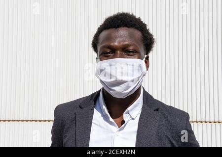 Jeune entrepreneur afro-américain positif en tenue formelle et de protection masque regardant la caméra en se tenant contre le mur dans la rue Banque D'Images