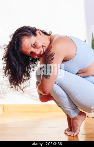 Vue latérale de la femme flexible dans les vêtements de sport pratiquant le yoga dans Pasasana sur les orteils à la maison tout en souriant à la caméra