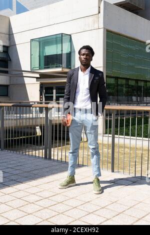 Corps complet de jeune homme d'affaires afro-américain dans élégant veste et jean tenant l'organisateur à la main et regardant Caméra à proximité d'un bâtiment urbain contemporain dans la ville de Barcelone Banque D'Images