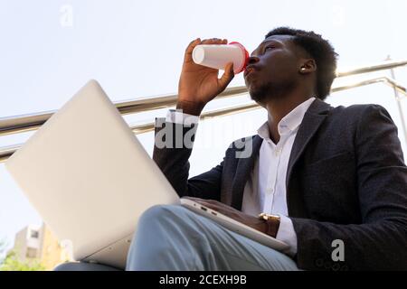 D'en-dessous de la vue de jeune homme d'affaires afro-américain confiant dans un costume formel, boire un café à emporter tout en étant assis en ville rue et navigation sur ordinateur portable Banque D'Images