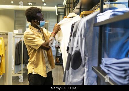 Vue latérale d'un homme afro-américain dans un masque médical debout dans le magasin de vêtements et de choisir t shirt pendant l'épidémie de coronavirus Banque D'Images