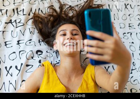 Angle élevé de la femme ravie couché sur le lit et prenant photo sur selfie tout en vous amusant pendant le week-end à la maison Banque D'Images