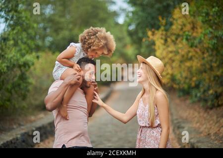 Jeune femme blonde gaie et mari afro-américain avec peu fille aux cheveux bouclés sur les épaules en appréciant la journée d'été ensemble pendant marche dans le parc Banque D'Images