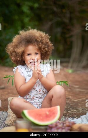 Gaie petite fille aux cheveux bouclés qui se claque les mains et regarde appareil-photo assis près d'un morceau de melon d'eau rouge mûr pendant pique-nique en été Banque D'Images