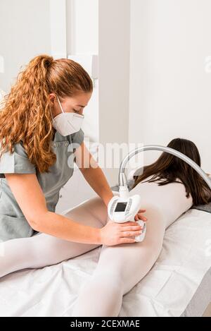 Masseuse dans le masque de protection faisant anti cellulite lipo massage à femme cliente vêtue de costume blanc pendant la procédure de soins du corps en salon de cosmétologie d'appareils Banque D'Images