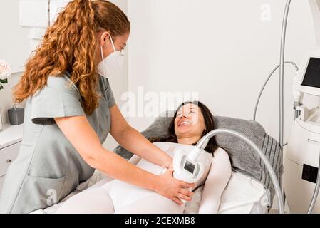 Masseuse dans le masque de protection faisant anti cellulite lipo massage à taille client femme vêtue d'un costume blanc lors des soins du corps procédure en salon de la cosmétologie de l'appareil Banque D'Images