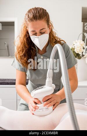 Masseuse dans le masque de protection faisant anti cellulite lipo massage à coupe courte non reconnaissable des hanches de la femme vêtues de blanc pendant procédure de soins du corps en salon de cosmétologie d'appareil Banque D'Images