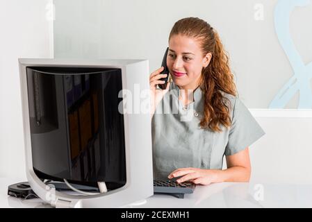 Une jeune réceptionniste souriante et agréable, en uniforme, parle au téléphone et de vérifier les informations sur l'ordinateur tout en étant assis sur le bureau bureau de clinique contemporain Banque D'Images
