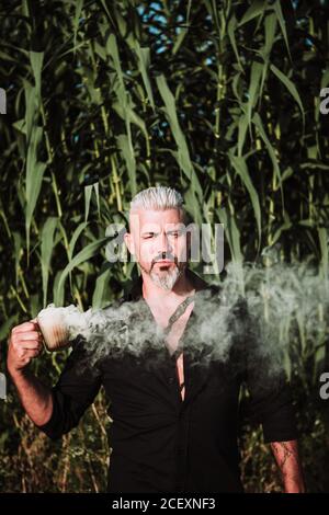 Homme aux cheveux gris brutaux avec barbe et moustache fumez la bombe et regardez la caméra en vous tenant debout contre de grands plants de maïs au champ Banque D'Images