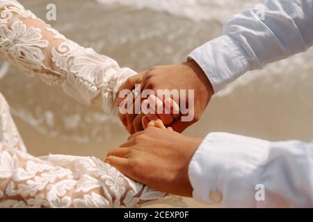 Vue latérale de Crop anonyme couple récemment marié dans élégant vêtements de mariage blancs tenant les mains tout en se tenant contre la mer Banque D'Images