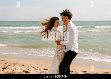 Vue latérale de jeune mariée gaie et marié dans l'élégant costumes de mariage debout s'embrassant les uns les autres avec les yeux fermés plage de sable près de la mer et de s'amuser Banque D'Images