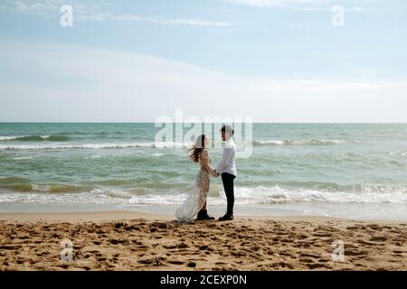 Vue latérale sur le marié et la mariée dans de beaux vêtements de mariage debout sur le sable près de la mer agitant pendant le voyage romantique tenue les mains et en regardant les uns les autres souriant Banque D'Images