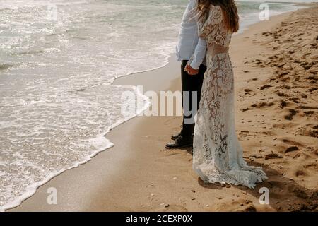 Vue latérale de la petite mariée et marié méconnaissable dans beau vêtements de mariage debout sur le sable près de la mer agitant pendant romantique voyage Banque D'Images